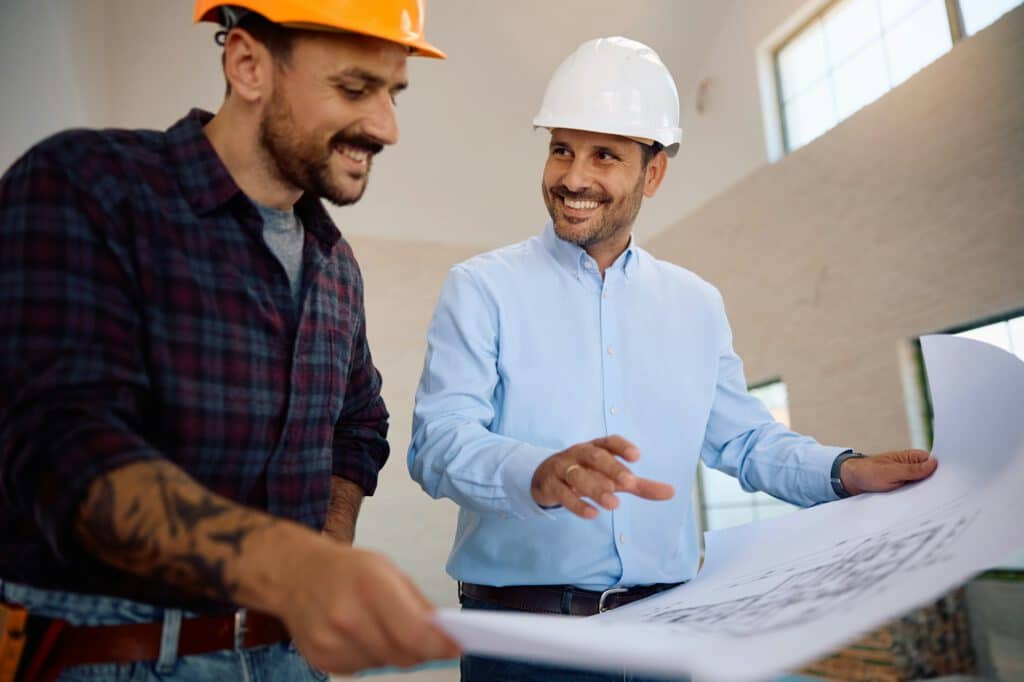 Happy housing project manager examining blueprints with a worker at construction site.